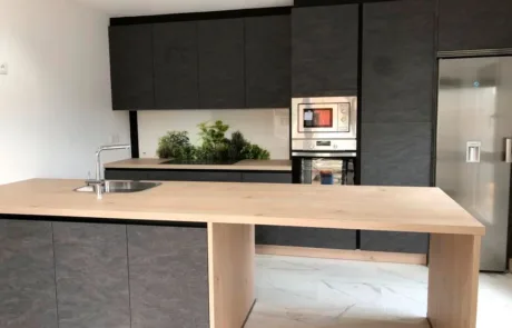 Cocina minimalista con isla central de madera clara y gabinetes oscuros texturizados. La cocina incluye un fregadero bajo la ventana, electrodomésticos modernos empotrados y una pared decorativa con plantas. El espacio es luminoso y combina elementos naturales con un diseño contemporáneo.